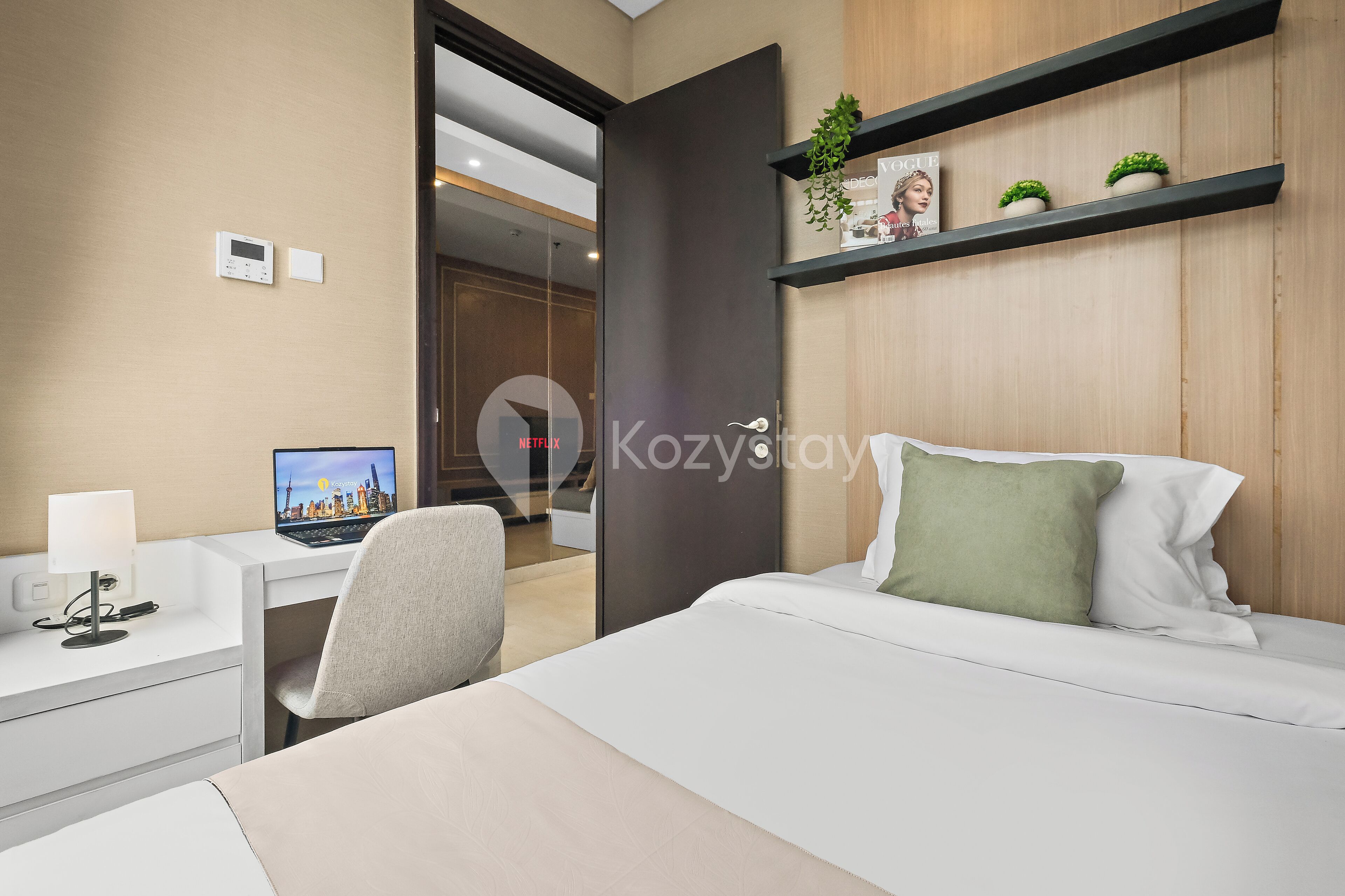 Une chambre d’hôtel moderne équipée d’un lit, d’un bureau avec un ordinateur portable, d’une chaise et d’une étagère avec des plantes.