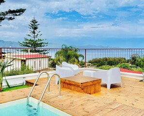 A wooden deck with a pool, white chairs, and a view of the sea and mountains. - Panoramic Villa with Infinity Pool – Relax and Comfort in Sicily (Sicilia)