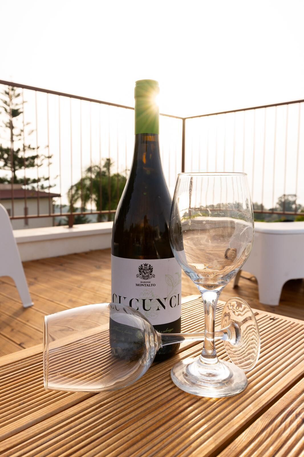 A bottle of Barone Montalto wine and an empty wine glass on a wooden table with a view of palm trees and a railing in the background.
