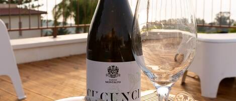 A bottle of Barone Montalto wine and an empty wine glass on a wooden table with a view of palm trees and a railing in the background.
