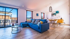 Ein modernes Wohnzimmer mit einem blauen Sofa, einem Essbereich mit einem Holztisch und großen Fenstern mit Blick ins Freie.