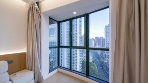 A modern hotel room with a large window, a bed, a bedside table, and a view of high-rise buildings.