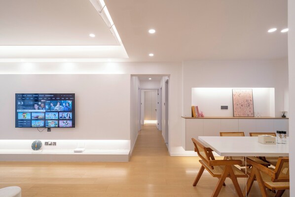A modern hotel room with a flat-screen TV mounted on the wall, a wooden floor, a white dining table with chairs, and a hallway leading to another room.