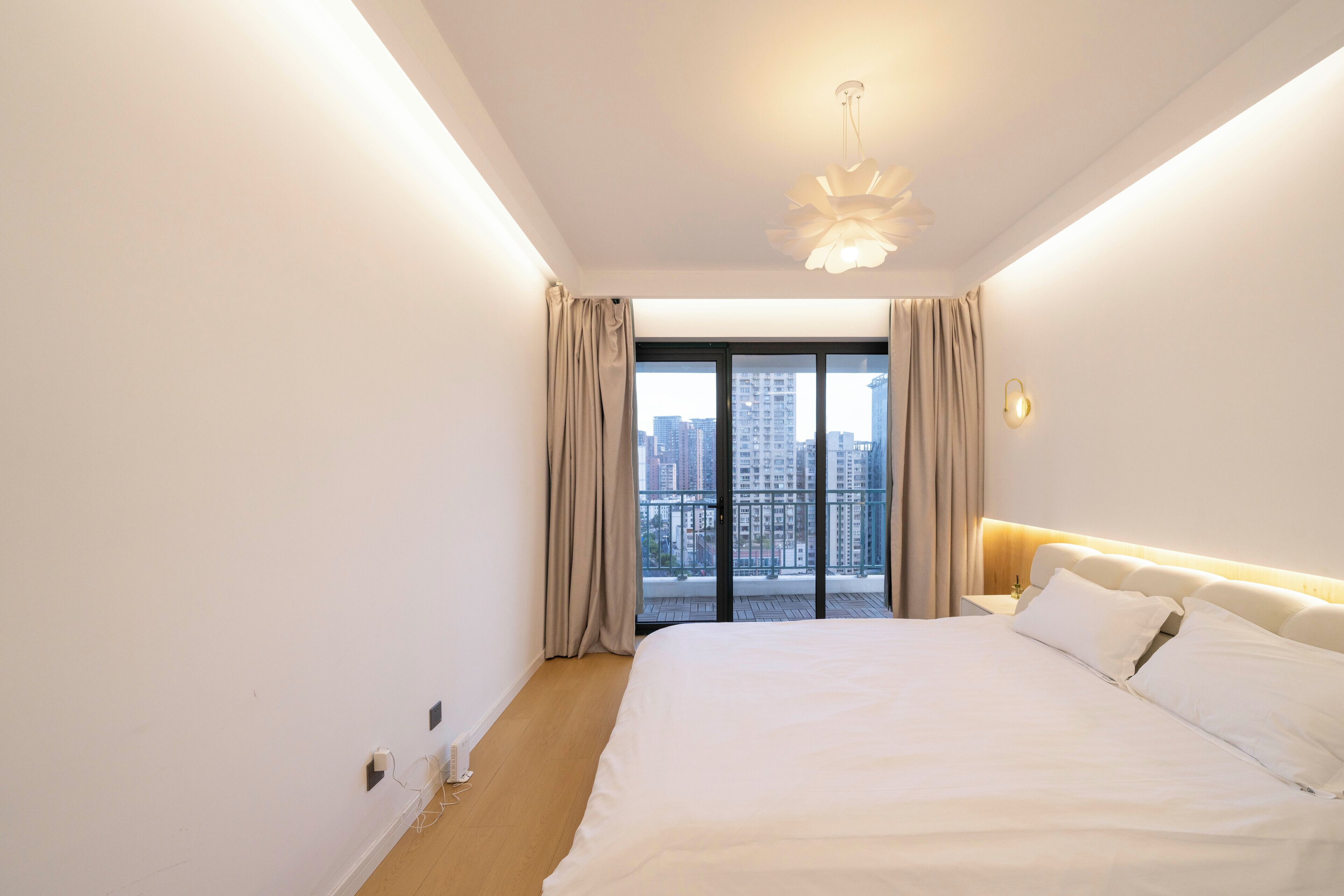 A hotel room with a large bed, a nightstand, a modern light fixture, and a view of a cityscape through a balcony with curtains.