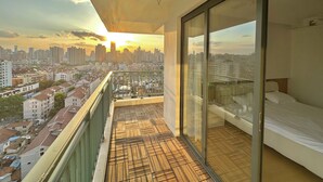 A balcony with a city view at sunset, a bed with a white mattress, and a wooden floor.