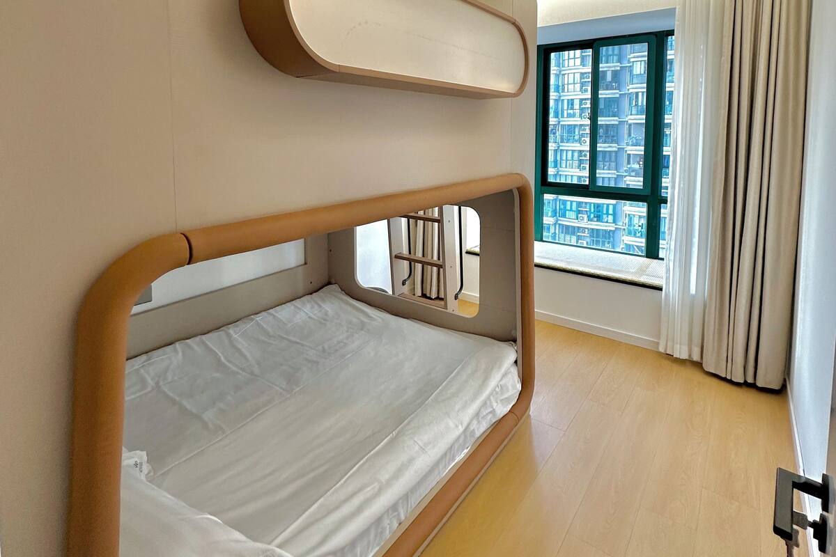 Un dormitorio moderno con una cama litera, un ventanal con vistas a edificios y una lámpara de techo.