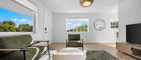 A modern living room with a sofa, a round table, a television, and a large window offering a view of greenery.