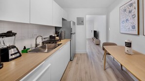 Ett modernt kök med vita skåp, en diskho i rostfritt stål, en mixer och ett kylskåp. Det finns ett träbord med en stol och ett inramat konstverk på väggen.