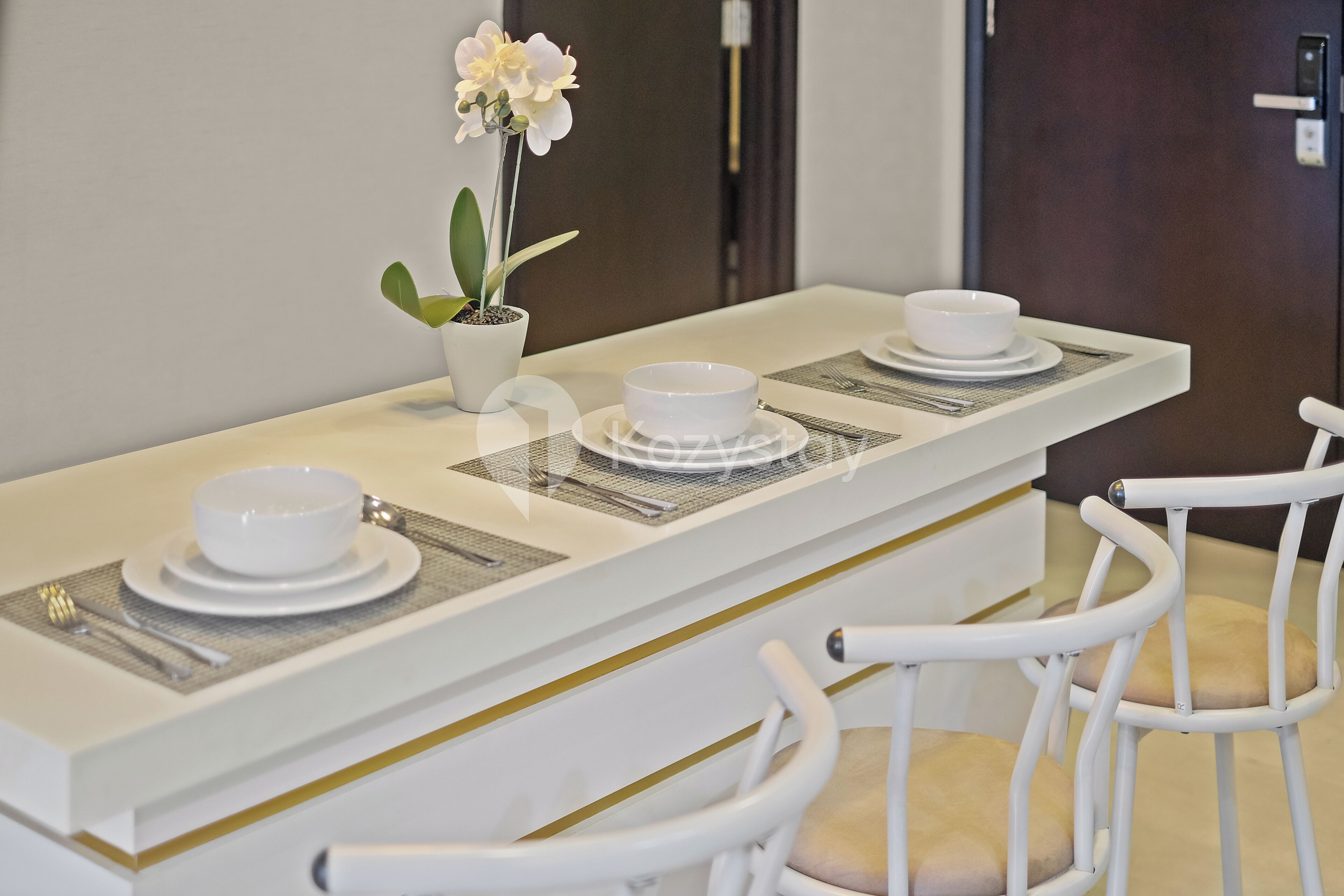 A dining area with a white table set for two, including plates, bowls, and cutlery. There is a potted orchid on the table.