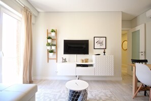 Una sala moderna con un televisor de pantalla plana montado en un mueble blanco, una mesita redonda pequeña, un sofá y un comedor con una silla.