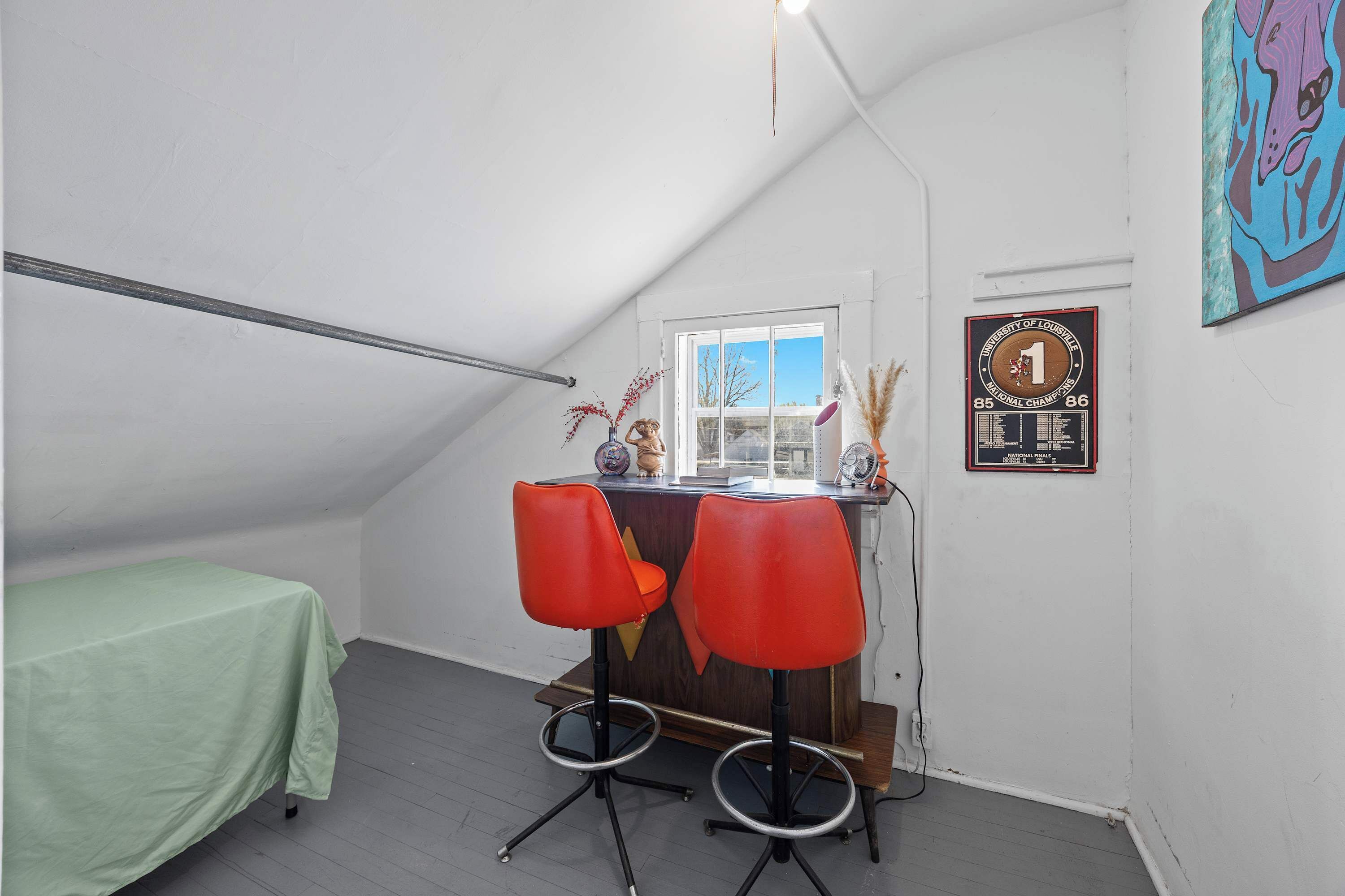 Una habitación ático pequeña y triangular con una cama individual, un escritorio y dos sillas rojas, una ventana y un certificado enmarcado en la pared.