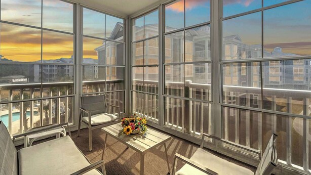 A sunlit balcony with large windows, a chair, a small table, and a floral arrangement. - Magnolia Pointe Unit 301 (Myrtle Beach)