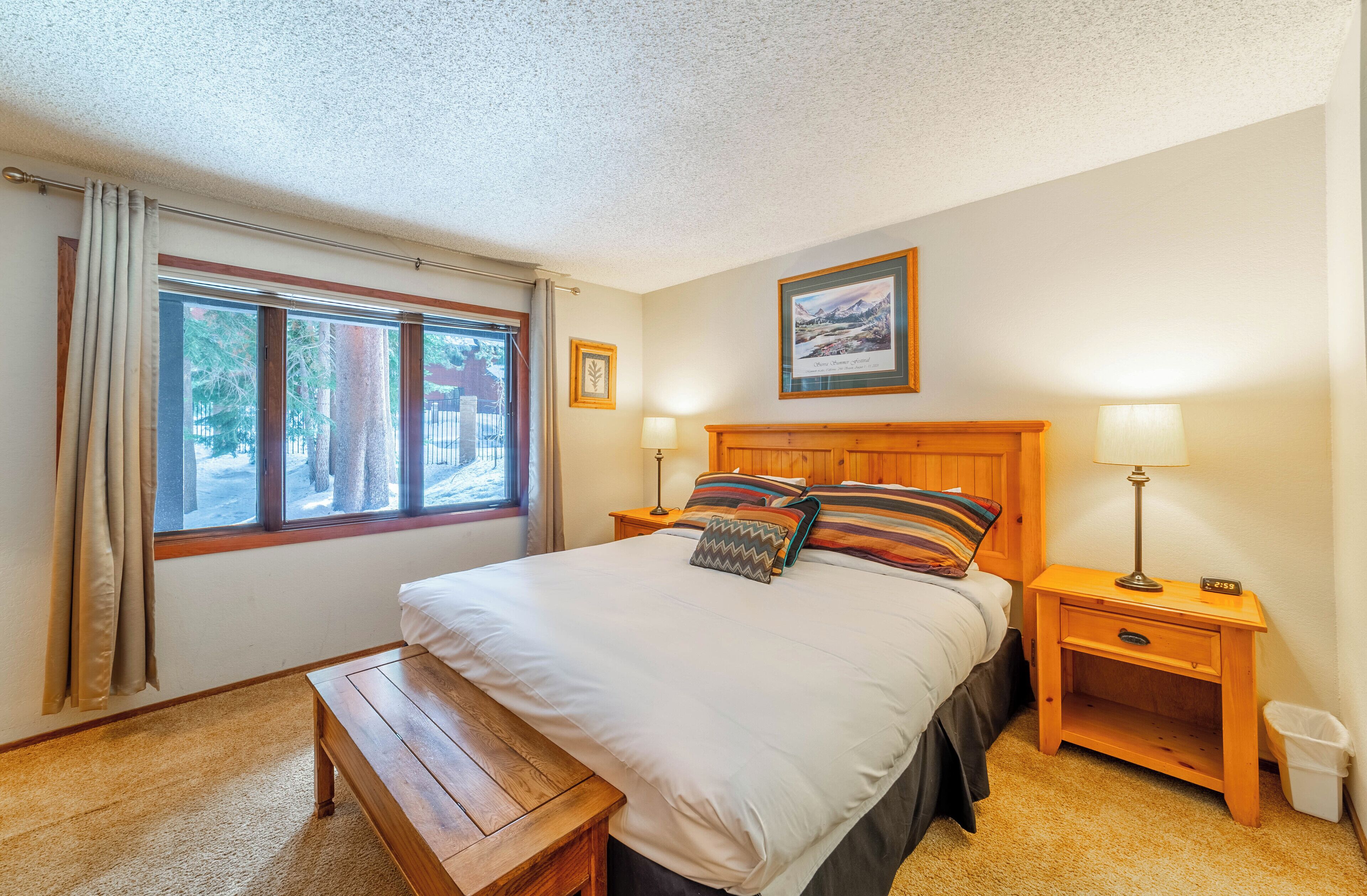 A bedroom with a large bed, two bedside tables, a bench, a window with curtains, and a framed picture on the wall.