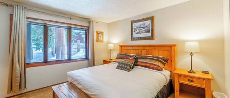 A bedroom with a large bed, two bedside tables, a bench, a window with curtains, and a framed picture on the wall.