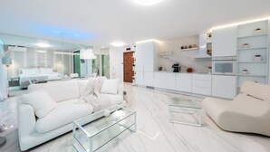 A modern, well-lit living room with a sofa, coffee table, and kitchen area with white cabinetry and marble flooring. - Emirates White Sensation (Sofia)