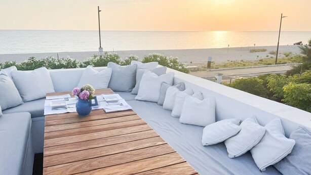 A sunlit outdoor seating area with a wooden table and cushions, overlooking a beach at sunset. - Elegant beachfront villa with jacuzzi, garden, and panoramic terrace. (Montignano-Marzocca)