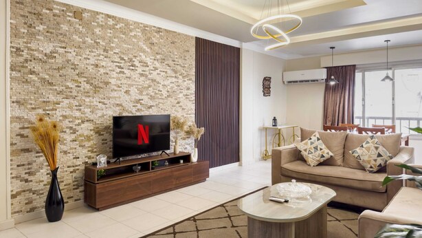 A modern living room with a brick-patterned wall, a flat-screen TV, a wooden entertainment unit, a beige sofa with patterned cushions, and a glass coffee table. - Xuru Stays I The Hospitable 2bdr Apt. 5 min From CAI Airport (Cairo)