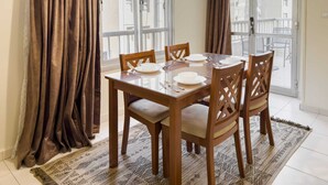 A dining area with a wooden table set for four, surrounded by chairs, with a patterned rug on the floor and a view of the outside through large windows. - Xuru Stays I The Hospitable 2bdr Apt. 5 min From CAI Airport (Cairo)