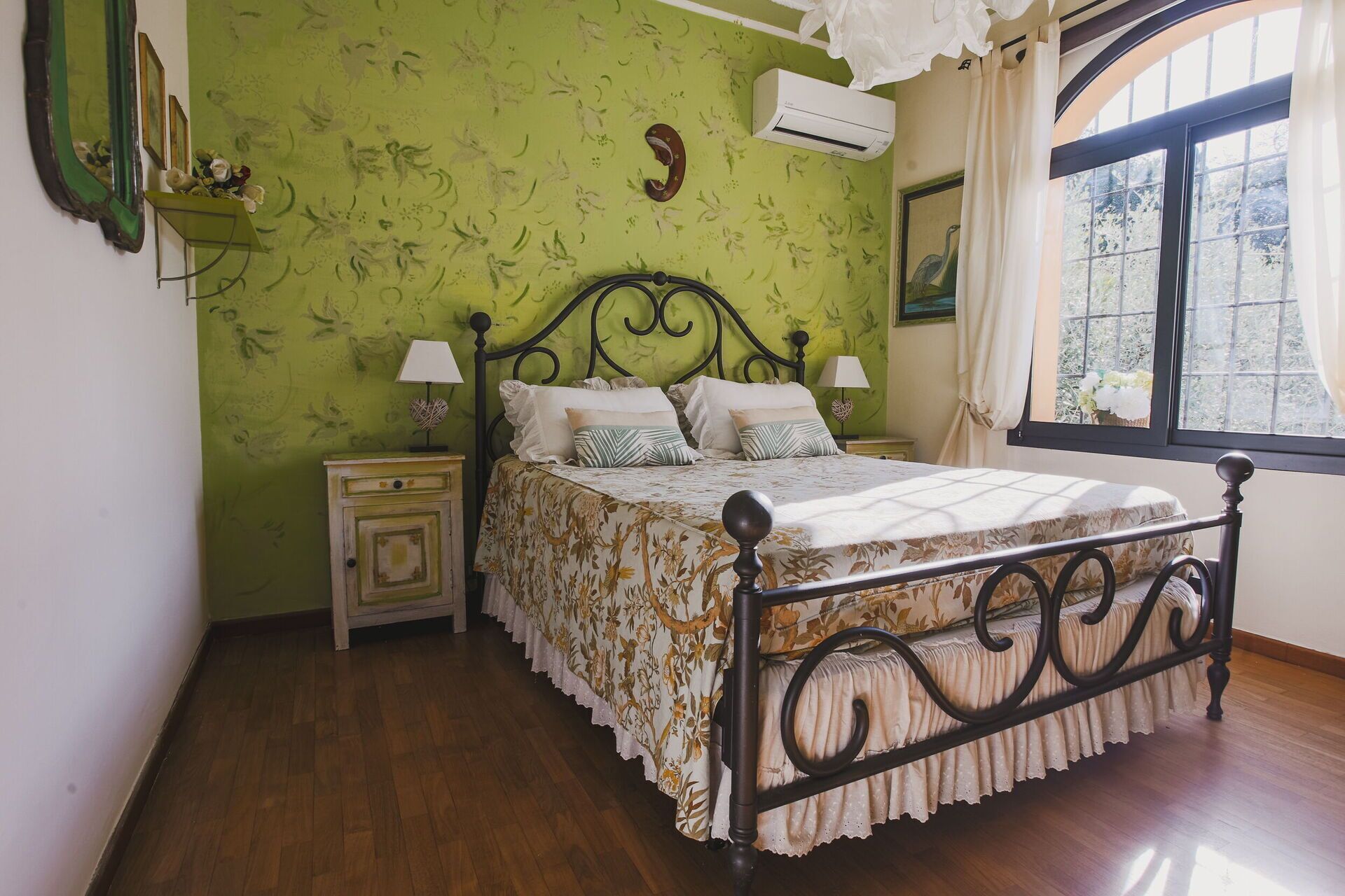 A bedroom with a floral bedspread, a wooden floor, a green wall with a floral pattern, a nightstand, a bedside table, and a window with curtains.