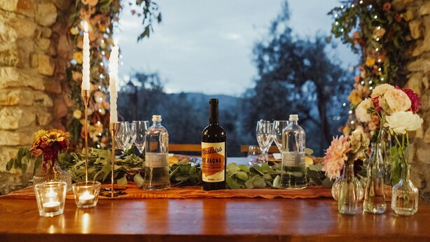 Et bord dækket til et måltid med en vinflaske, glas og blomsterarrangementer.