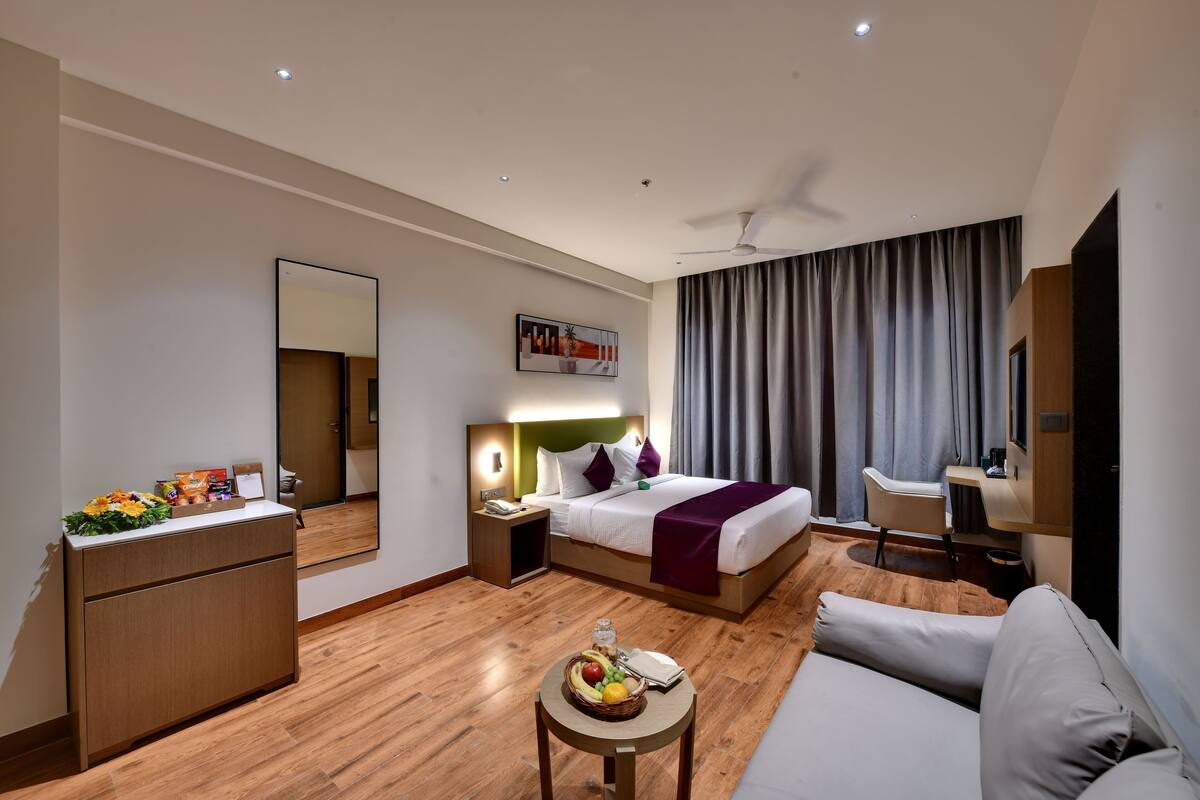 A modern hotel room with a bed, a desk, a sofa, a dining table with fruit, and a mirror.