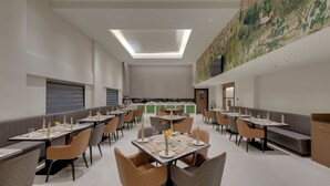 A spacious dining area with high ceilings, recessed lighting, and a large mural on the upper wall. Tables are set with white tablecloths, and there are multiple seating options including booths and chairs.