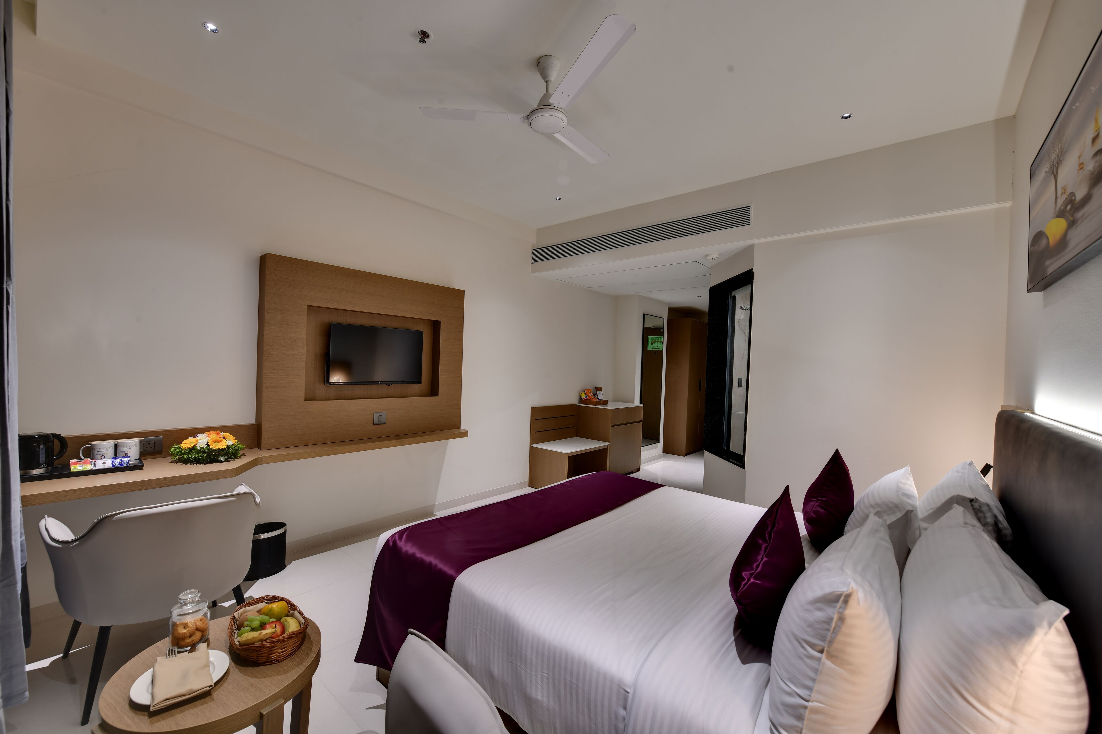 Habitación de hotel con cama, escritorio con televisor, una mesita con cesta de fruta y una silla.