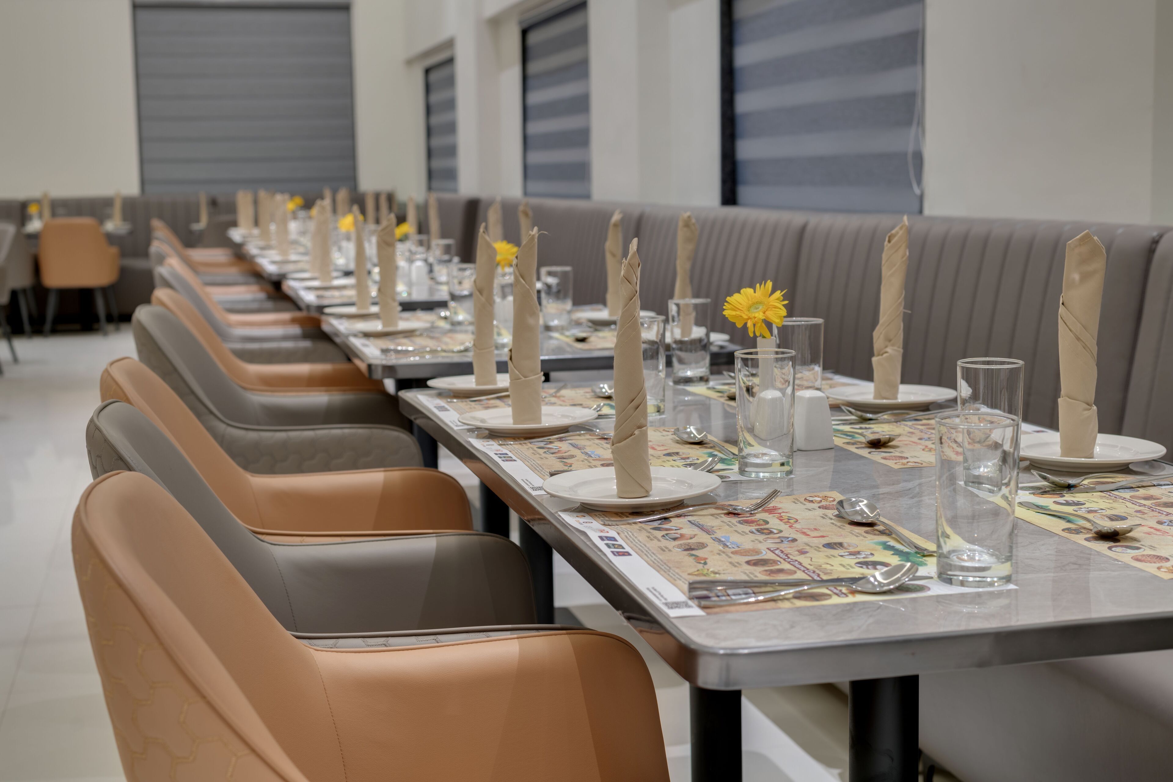 A dining area with multiple tables set for service, each with a vase of yellow flowers, tall candles, and clear glasses.