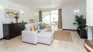 Un salon moderne avec un canapé blanc, un meuble en bois foncé, un téléviseur à écran plat et une grande fenêtre avec des rideaux.