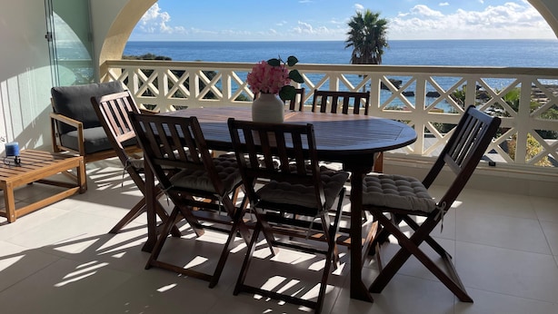 Un balcon avec une table ronde et des chaises, un vase rempli de fleurs et une vue sur l’océan.