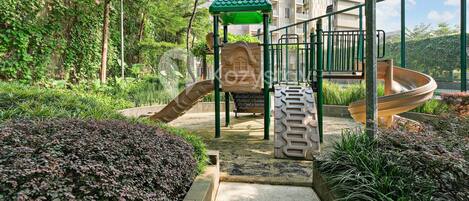 A playground with a slide and climbing structures, surrounded by greenery and a building in the background.
