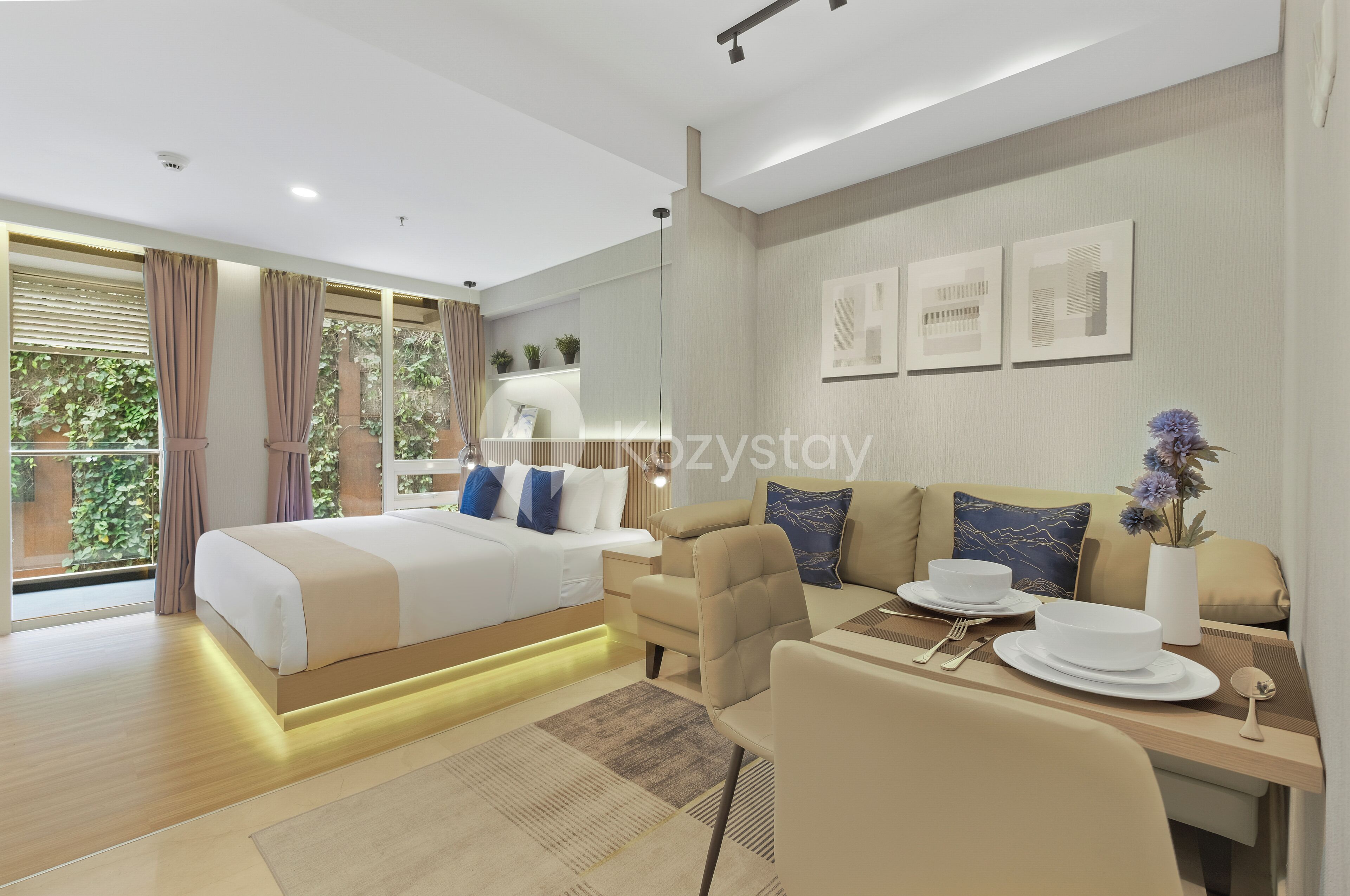 A modern hotel room with a large bed, a sofa, a dining table with plates and cutlery, and a view of greenery outside.