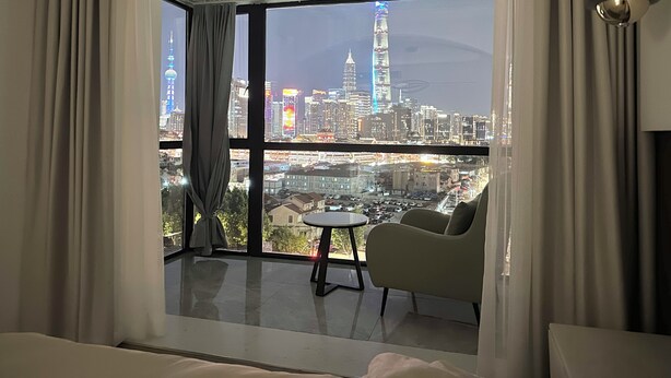 A modern hotel room with a large balcony, a round table, and a comfortable chair, offering a view of a city skyline at night. - ZHome-Sun Garden-Amazing view (Shanghai)