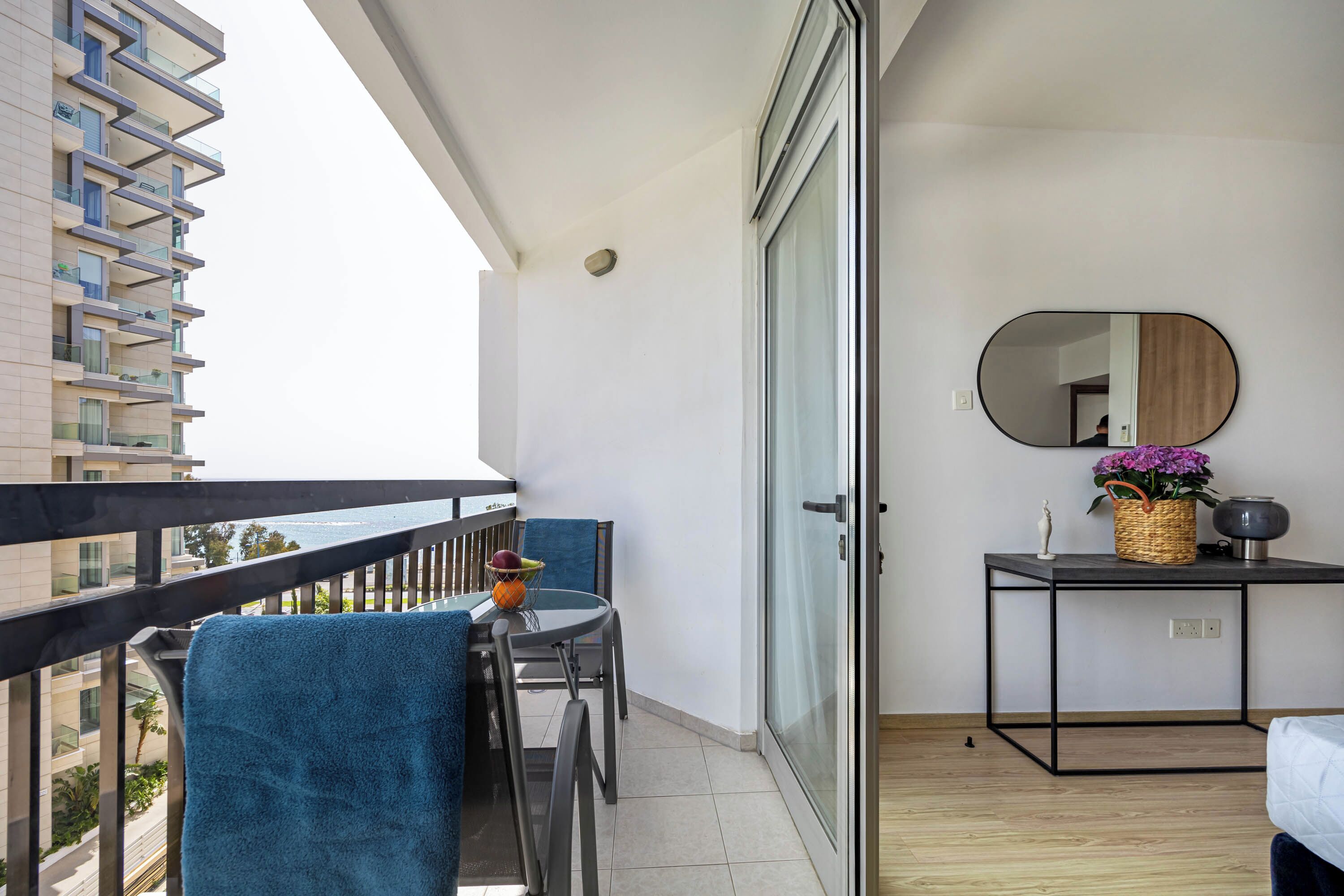 A balcony with a table, chairs, a small table with a basket and vase, and a sliding glass door leading to a bedroom.