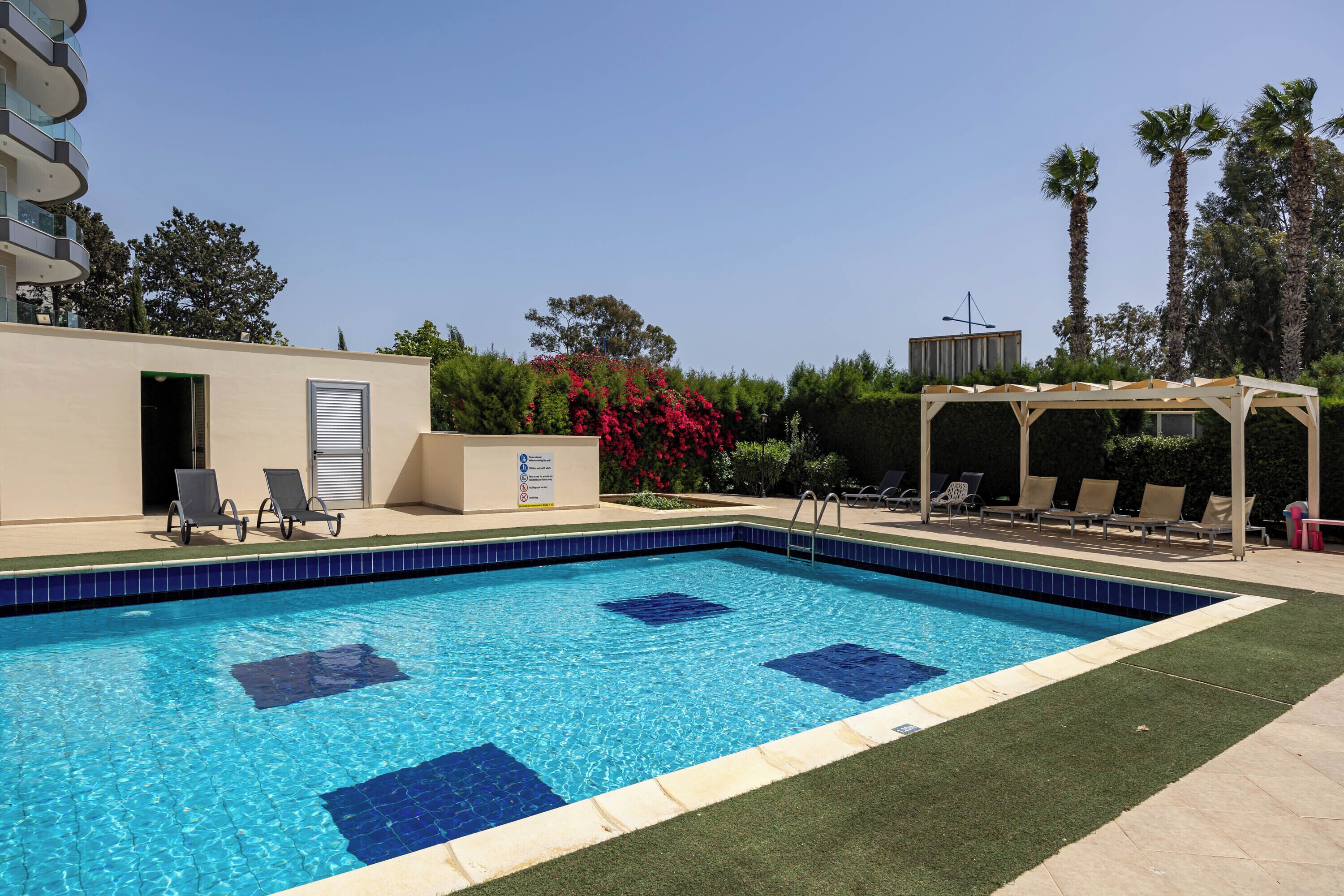 Piscina rectangular con azulejos azules, rodeada de césped verde y una pérgola con sillas.