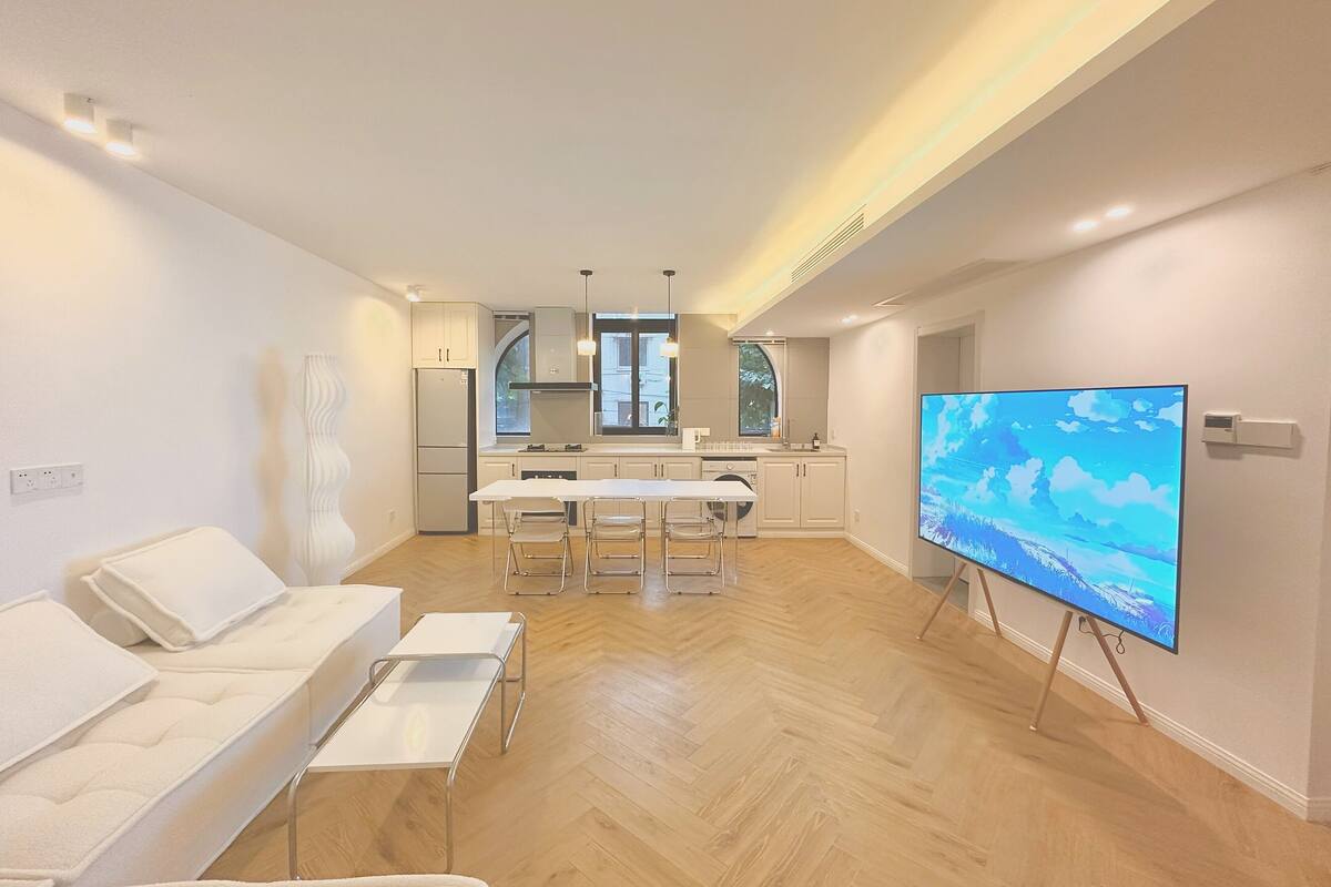 A modern living room with a sofa, a TV on a stand, and a kitchen in the background.