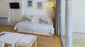 A room with a white sofa, two pillows with nautical designs, a small table, a television on the wall, and a large window with curtains. - Apartment -450m from the beach- swimming pools (La Baule-Escoublac)