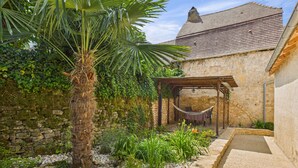 A stone building with a tiled roof, a hammock under a wooden pergola, and a variety of plants and shrubs.