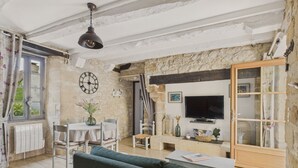 A rustic room with exposed stone walls, a wooden ceiling, a dining area with a table and chairs, a sofa, a television, and a bookshelf.