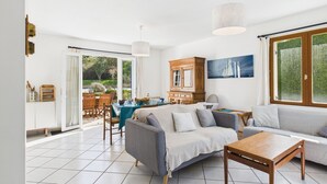 A spacious living area with a sofa, a coffee table, and a dining table set for four. There is a large window providing a view of the outdoors. - House for 6 by the sea - Garden and swimming pool (Pléneuf-Val-André)