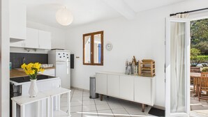 A modern kitchen with white cabinets, a white fridge, and a stove. There is a window with a view of greenery outside. - House for 6 by the sea - Garden and swimming pool (Pléneuf-Val-André)
