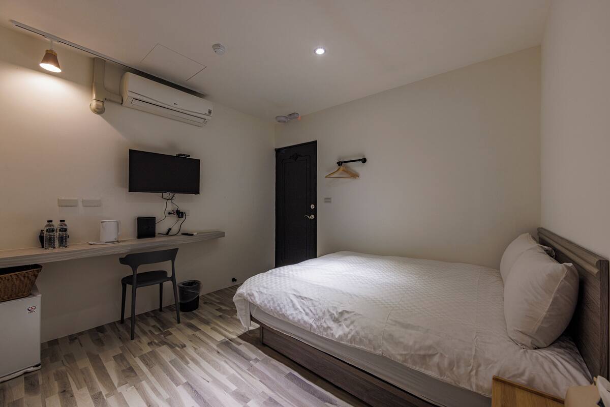 A bedroom with a bed, a desk with a chair, a TV, and a door.
