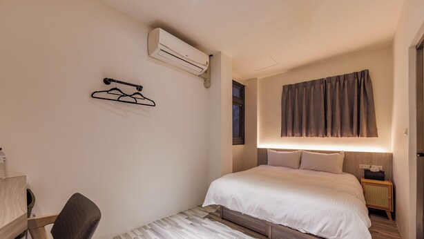 Ein Hotelzimmer mit Bett, Sessel, kleinem Tisch, Klimaanlage, Wandhänger, Nachttisch mit Lampe und Fenster mit Vorhängen.