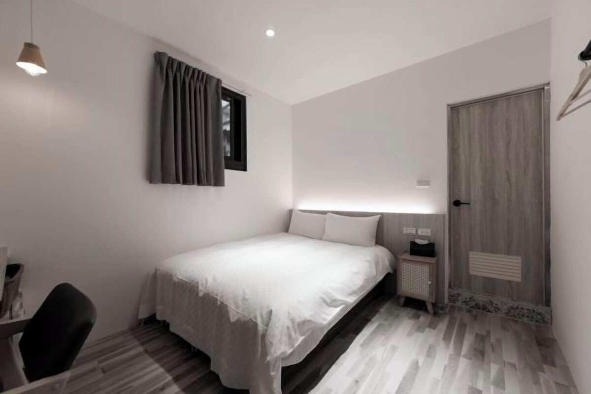 A modern hotel room with a neatly made bed, a bedside table with a lamp, a chair, and a wardrobe with a mirror.