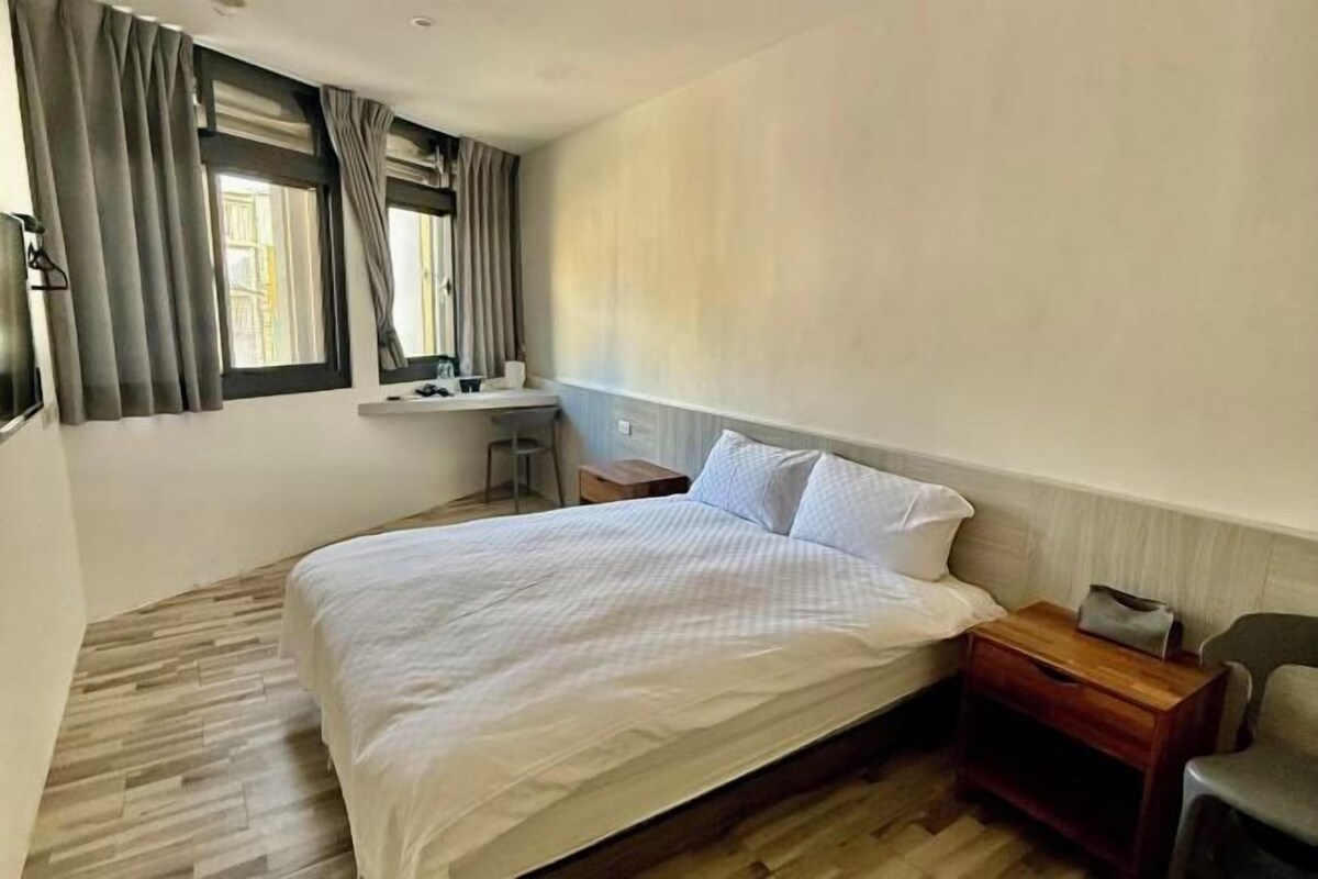 A bedroom with a bed, a bedside table, a chair, a desk, and a window with curtains.