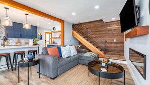 Un espacio habitable moderno con un sofá seccional gris, una mesa redonda de centro y una escalera con barandillas de madera.