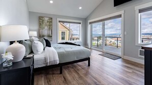 Un dormitorio moderno con una cama grande, una mesita de noche, una lámpara y vistas a la ciudad a través de amplios ventanales.