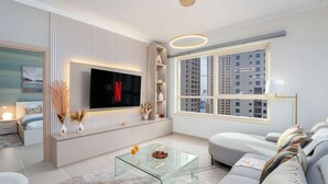A modern living room with a flat-screen TV mounted on a wall, a glass coffee table, a sofa, and a large window with a city view. - Ethereal 1 BR in JBR (Dubai)