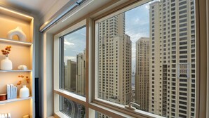 A room with a large window overlooking high-rise buildings, a shelf with decorative items, and a view of the cityscape. - Ethereal 1 BR in JBR (Dubai)
