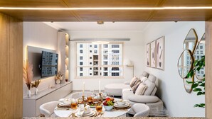 A modern living room with a dining area, a sofa, a TV mounted on the wall, and a large window with a view of other buildings. - Ethereal 1 BR in JBR (Dubai)